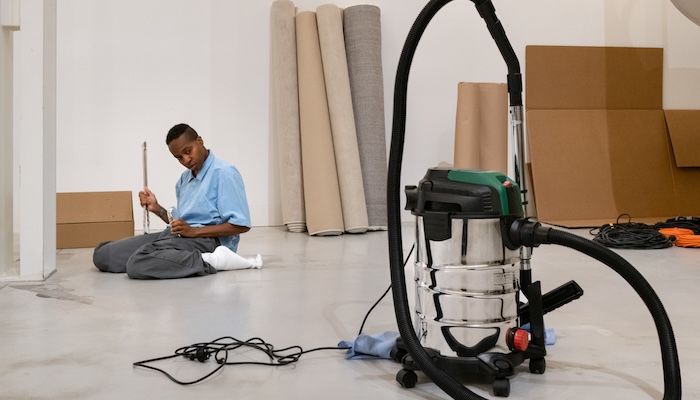A person sits in the background, staring at a large canister vacuum in the foreground.