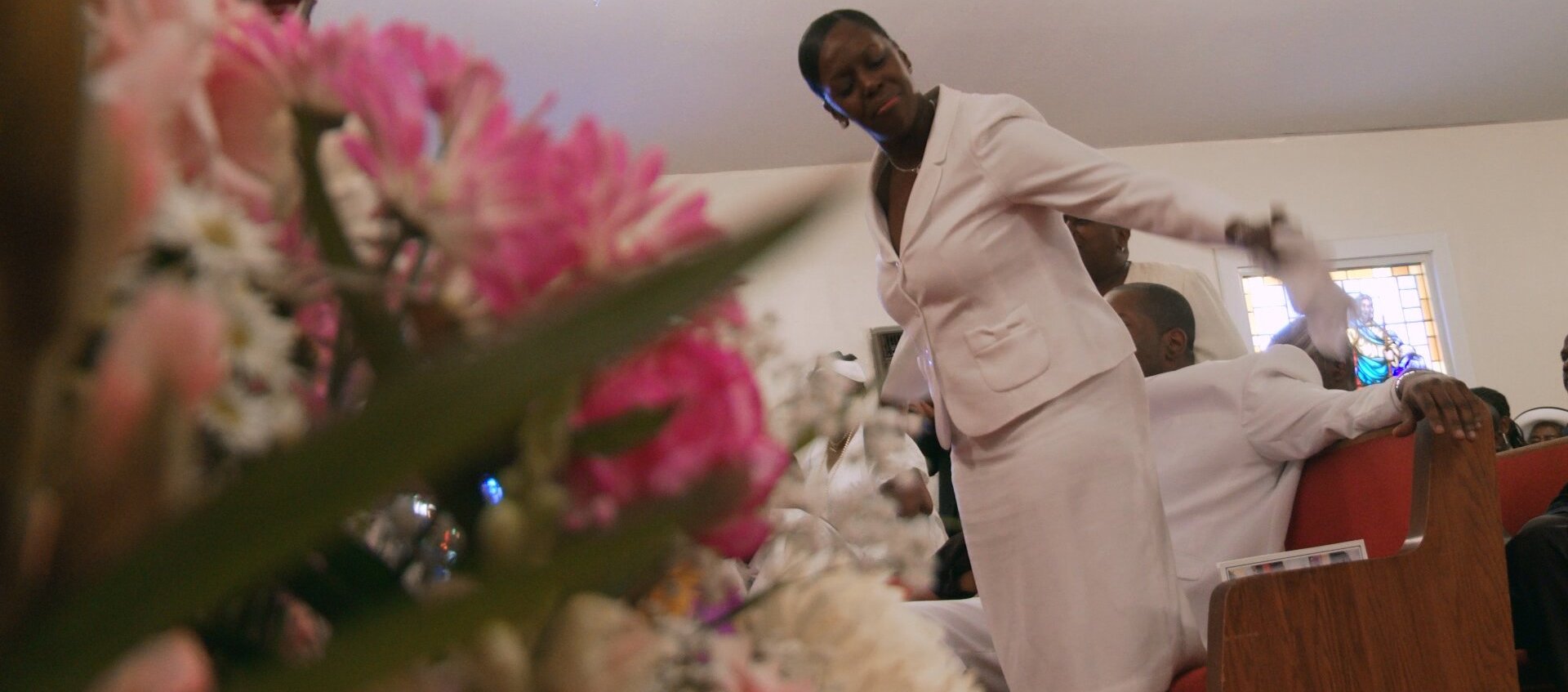 A Black feminine-presenting person is mid-movement as she attends a funeral at the Owens Funeral Home behind a close-up of a bundle of pink and white flowers.
