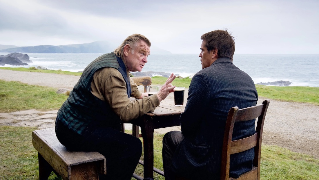 Two white men sit at a table outside with a mountanous coastal landscape in the background. They are engaged in a heated conversation with the man to the right pointing at the man to the left.