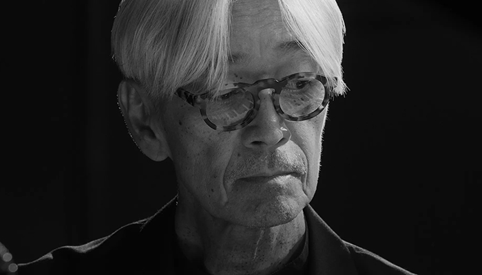 Headshot of Ryuichi Sakamoto, an older man with white hair and round glasses.