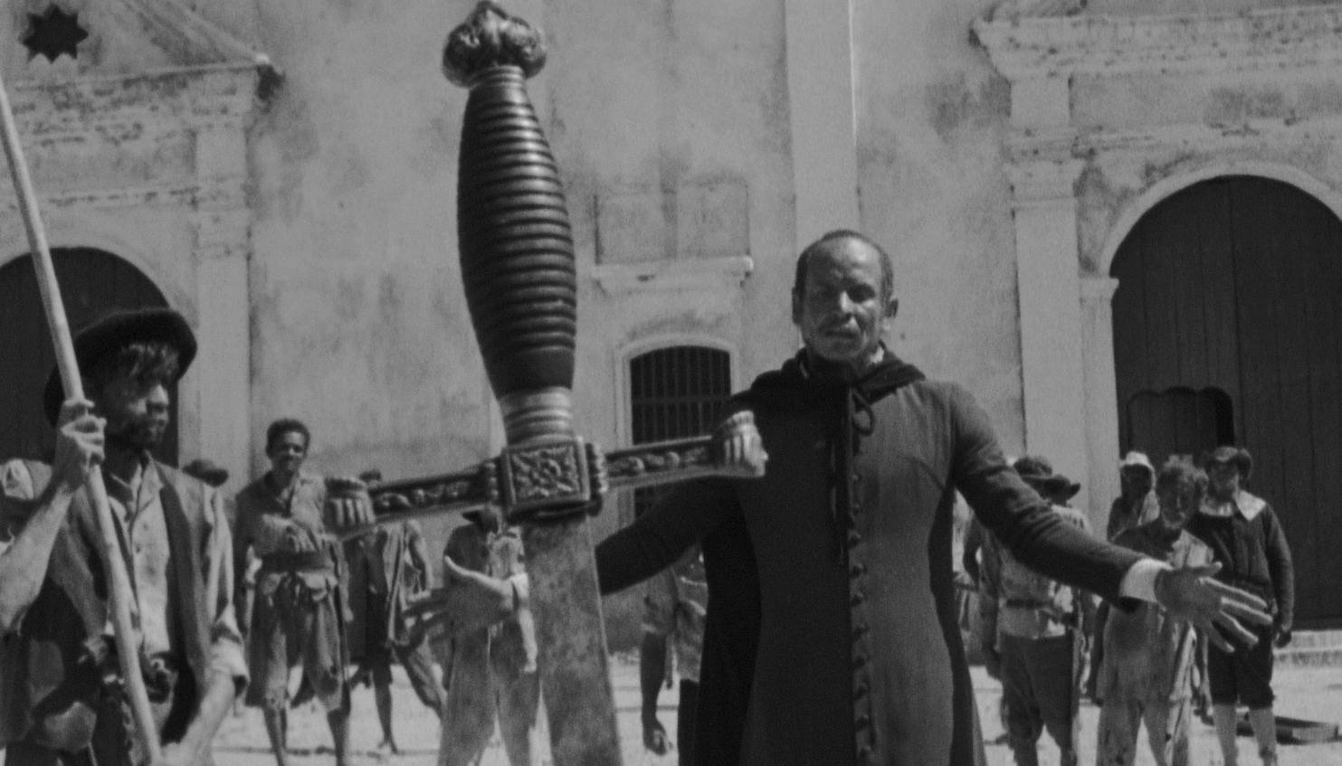 A man in religious garb stands with his hands out to his sides in front of a large stone building behind a giant sword stuck in the ground.