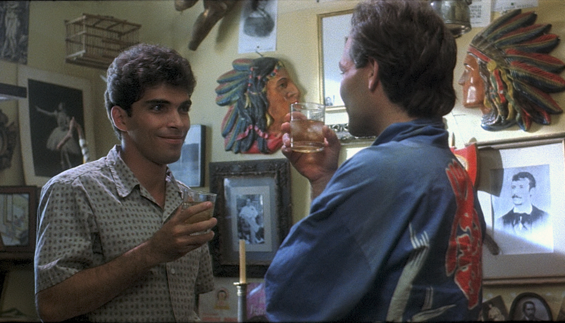 Two men stand facing eachother holding drinks in a room with many framed pictures on the wall.