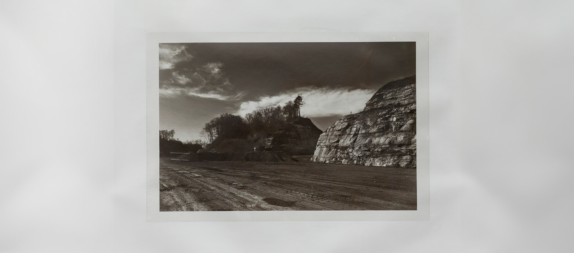 Framed black-and-white photo of a dirt road running diagonally across the composition with tall, partly tree-covered rock faces to the right. 