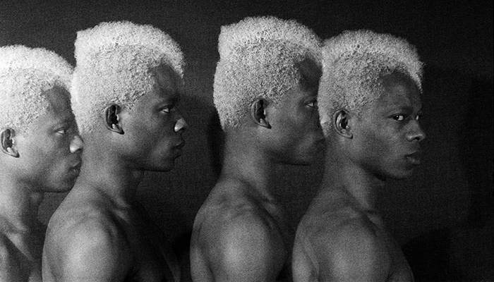 Photographic montage of repeated profile views of a young black male with white hair. In three he stares ahead; in the fourth he looks sideways at the camera.