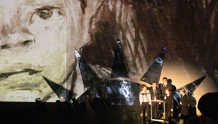 An image from the original production of Calling Hours, a large drawing of a child’s face is projected behind a group wearing suits and black dresses. 