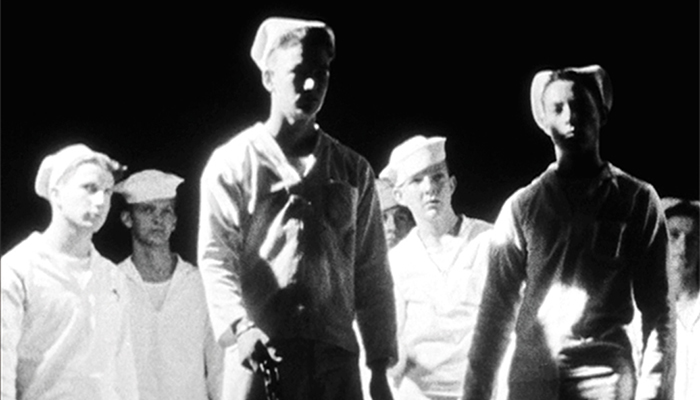 Black-and-white still of a group of young, white adults with short hair; they are all wearing white uniforms and sailor’s hats in front of a black background.