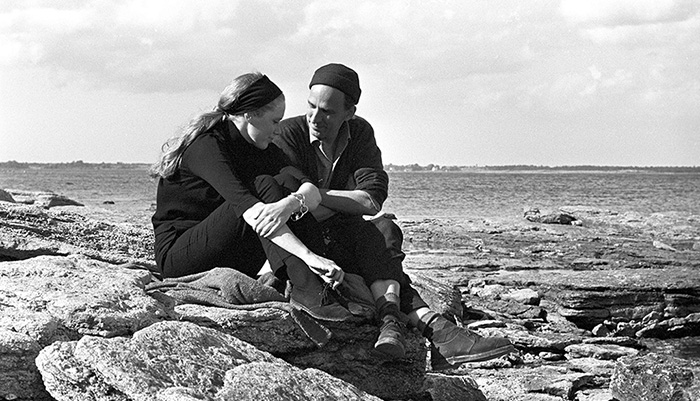 A black and white photo of a white man and a white woman sitting on a cliff by the sea.