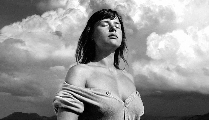 a black and white image in a sweater pulled down below her shoulders with eyes closed against a cloudy sky