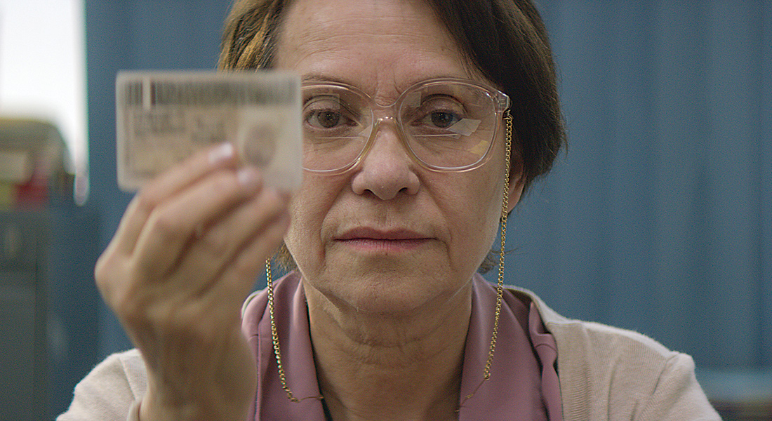 A middle-aged Latino woman in glasses seen in close up against a blue wall, facing the camera and holding an ID card up in front of her