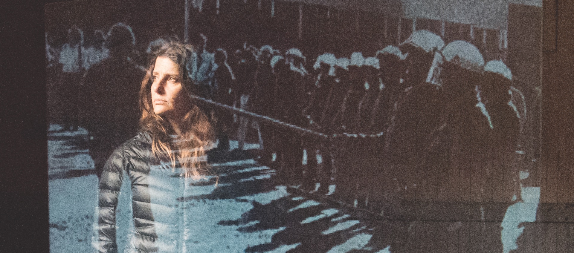Actress Mia Barron stands behind a transparent screen with an image of police in riot gear during performance of Lars Jan & Early Morning Opera's production of Joan Didion's The White Album