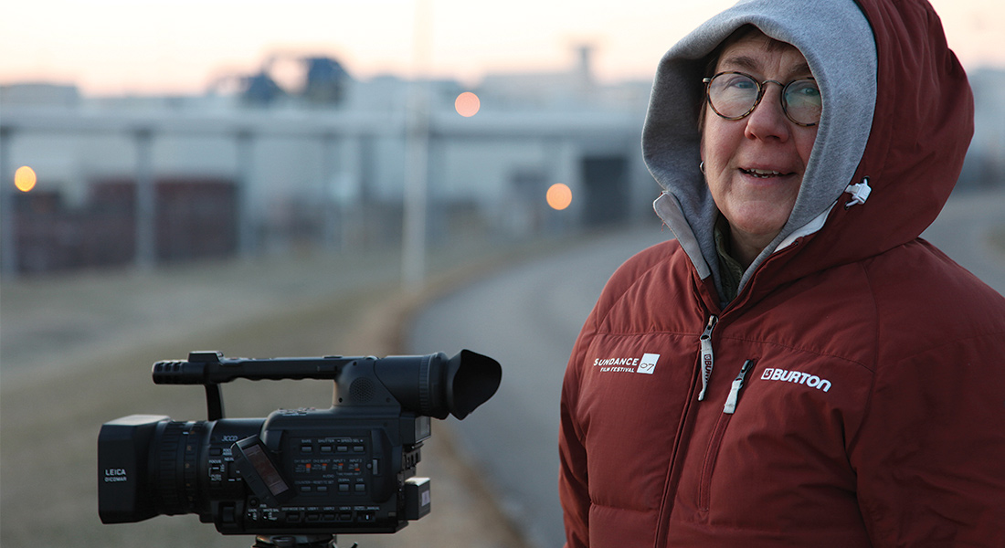 A photo of filmmaker Julia Reichert standing outside in the cold, wrapped in a red hooded jacket, with a digital movie camera