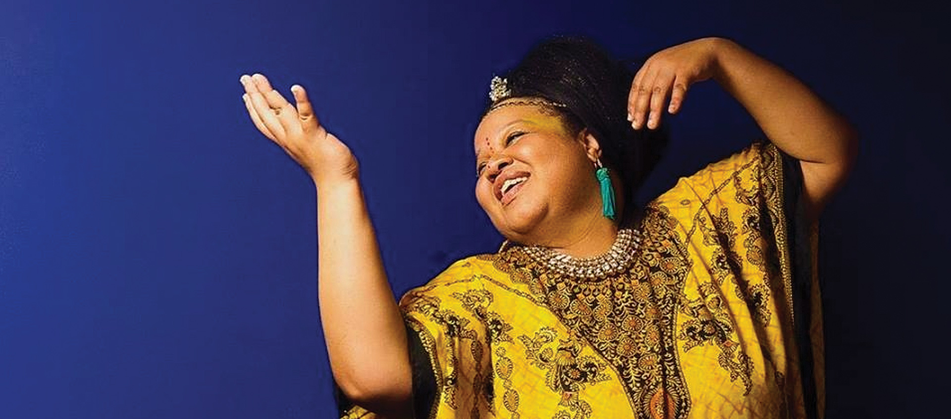 A picture of Chicago-based free jazz artist Angel Bat Dawid in a patterned bright yellow dress, smiling and looking to her right against a blue background