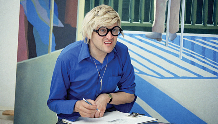 An image of British artist David Hockney sitting in front of one of his paintings in a scene from Jack Hazan's 1973 film A Bigger Splash