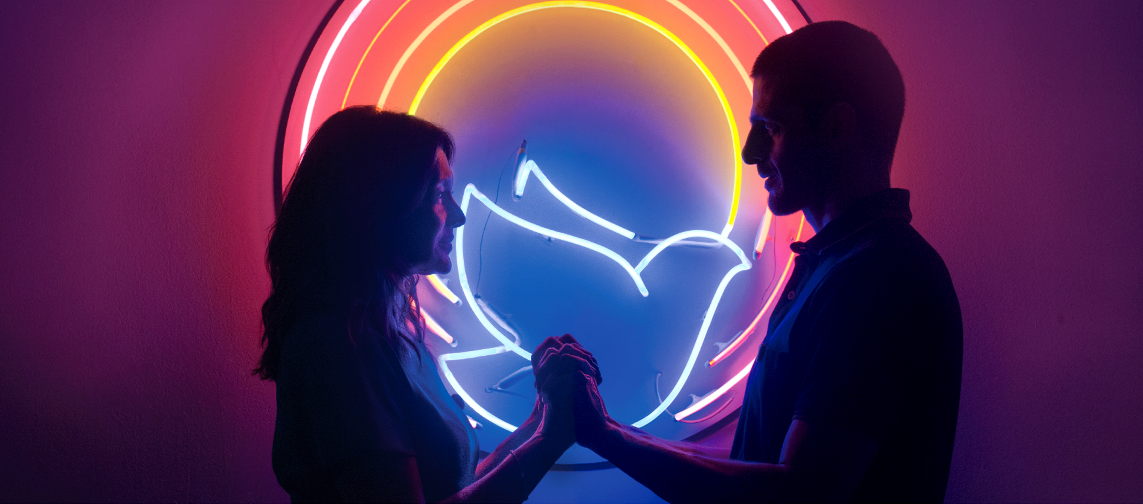 Two humans standing in front of a neon sign with a bird in the center