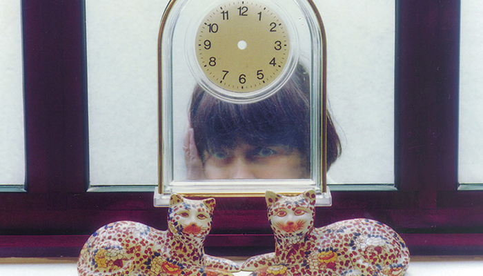 Filmmaker Agnés Varda seen from the cheeks up, standing behind a glass table clock and two decorative ceramic cats