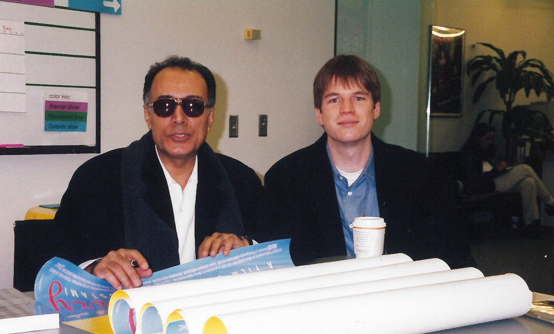 A photo of Iranian director Abbas Kiarostami sitting with Wexner Center Film/Video Director David Filipi behind a table with posters for Kiarostami's 1998 film A Taste of Cherry