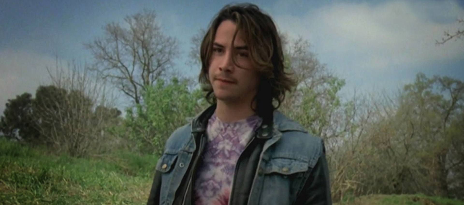 Color image of a young Keanu Reeves standing outdoors in front of grass and trees during the production of Tim Hunter's 1984 film River's Edge