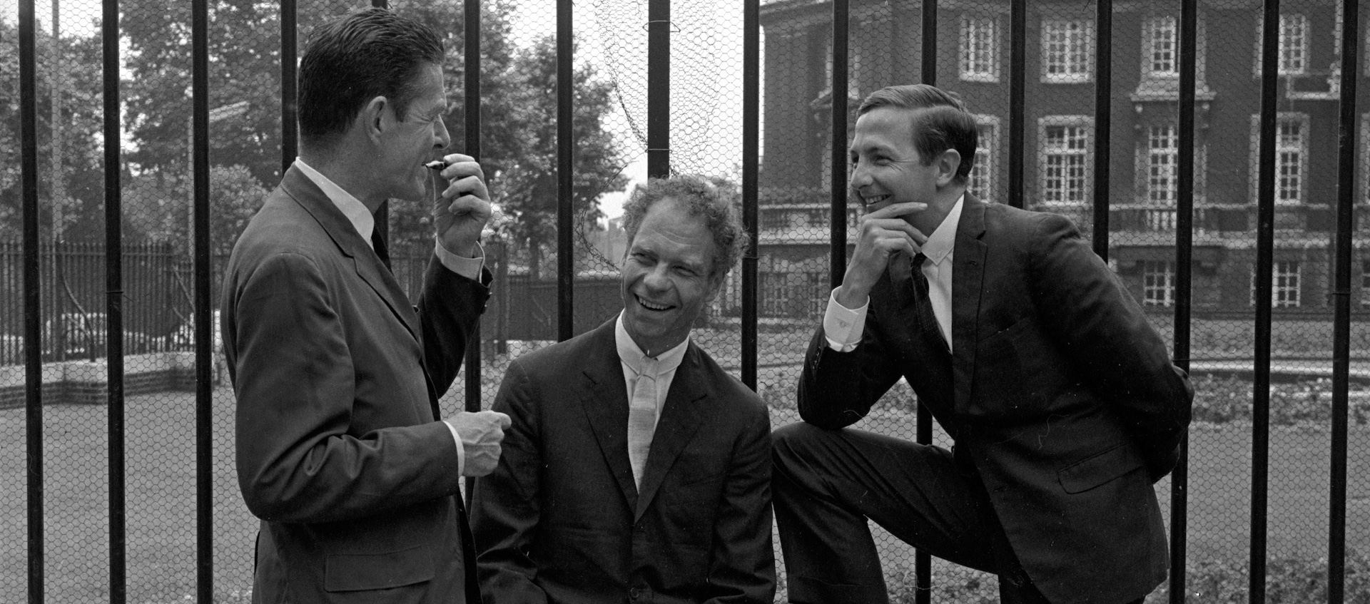 A black and white image of choreographer Merce Cunningham sitting against a tall iron fence, looking at the camera. He sits between composer John Cage and artist Robert Rauschenberg, who are both standing and facing each other