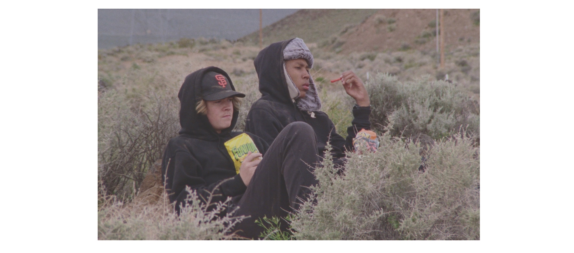 Two teenage boys in hats and coats sit in the California wilderness eating snack food in a scene from Stanya Kahn's 2020 short film No Go Backs