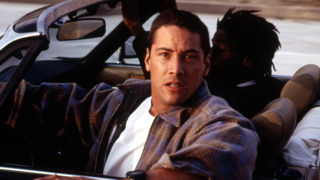 A color image of Keanu Reeves facing the camera while in the driver's seat of a car in a scene from Jan de Bont's 1994 film Speed
