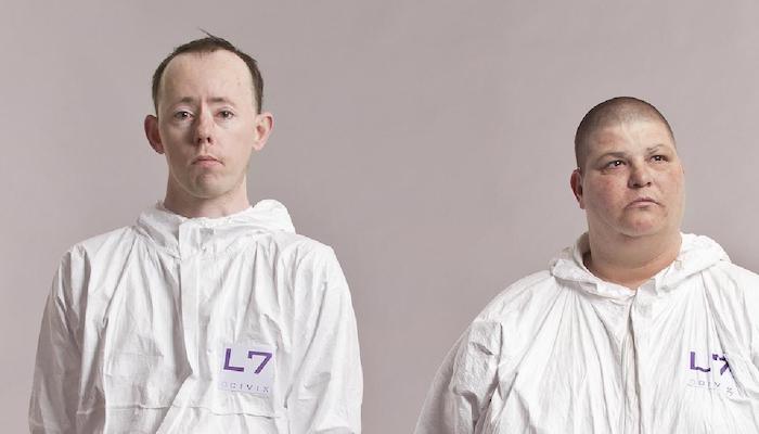 Back to Back Theatre company members Simon Laherty and Sonia Teuben in white hazmat outfits, seen standing shoulders up against a solid beige background