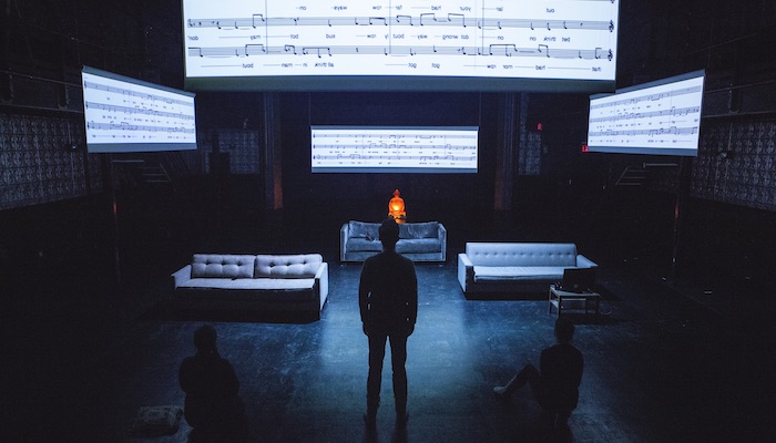 A man stands with his back to the camera on a darkened stage with individuals sitting on either side of him as he looks up at screens showing the notes in a music composition in a scene from Annie Dorsen's performance work Yesterday Tomorrow