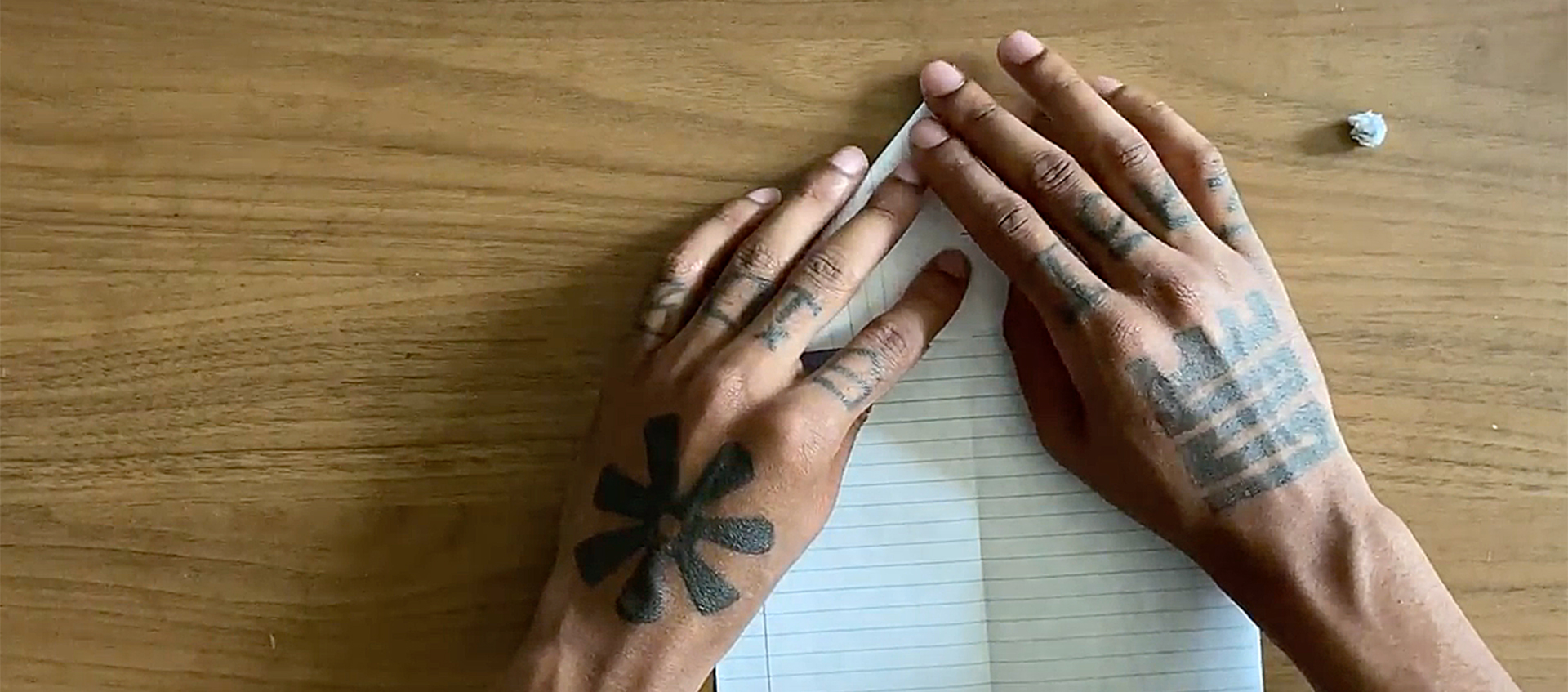 The tattooed hands of African American artist Hakim Callwood begin to fold a piece of lined paper on a wooden surface into a paper airplane