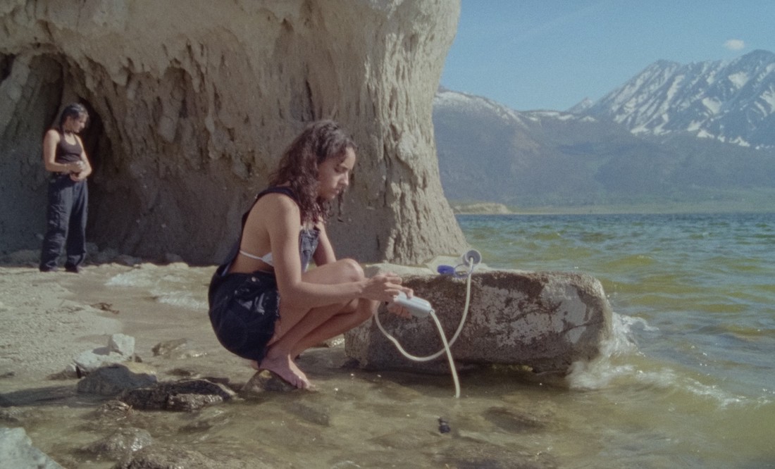 A teenage girl crouches by the shore, pumping sea water through a filter pump as another girls stands in the distance behind her in a scene from Stanya Kahn's No Go Backs