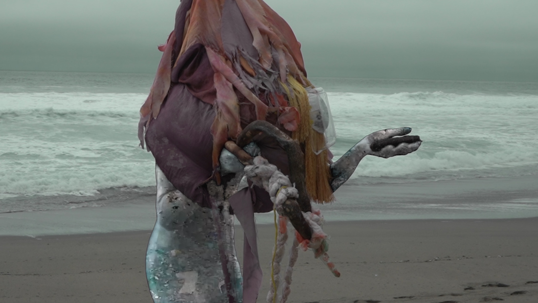 A figure on the beach appears to be tangled in plastic and trash from the waist up in a scene from Connie Zheng's video The Lonely Age (Part 1)