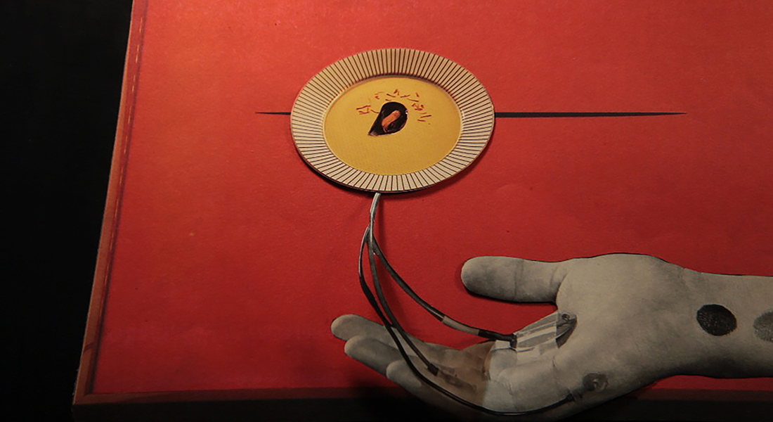 In this color collage, a still from the film, a hand is extended palm-up with wires coming out of it that connect to a bowl of soup.
