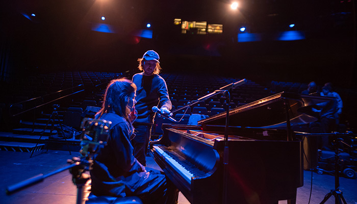 Erin Durant sits at a piano on stage as a crew person places a mic in front of her