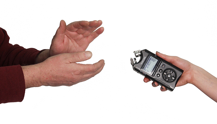 A person out of frame except for their hands claps on the left side while on the right, a hand with a small recorder in it extends toward the person clapping