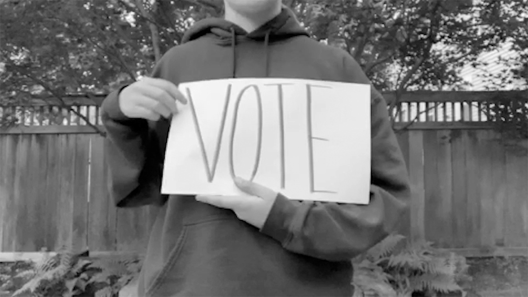 A person in a sweatshirt stands against a backyard fence and holds a sign with VOTE written on it.