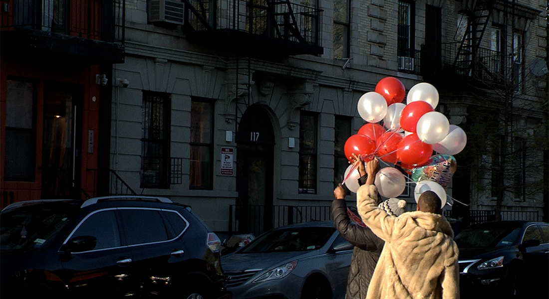 Two men hold up bunches of red and white balloons outside an old residential building in Brooklyn