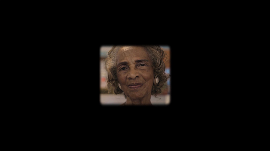 An elderly African American woman's face smiling in a small square frame at the center of the image, surrounded by black