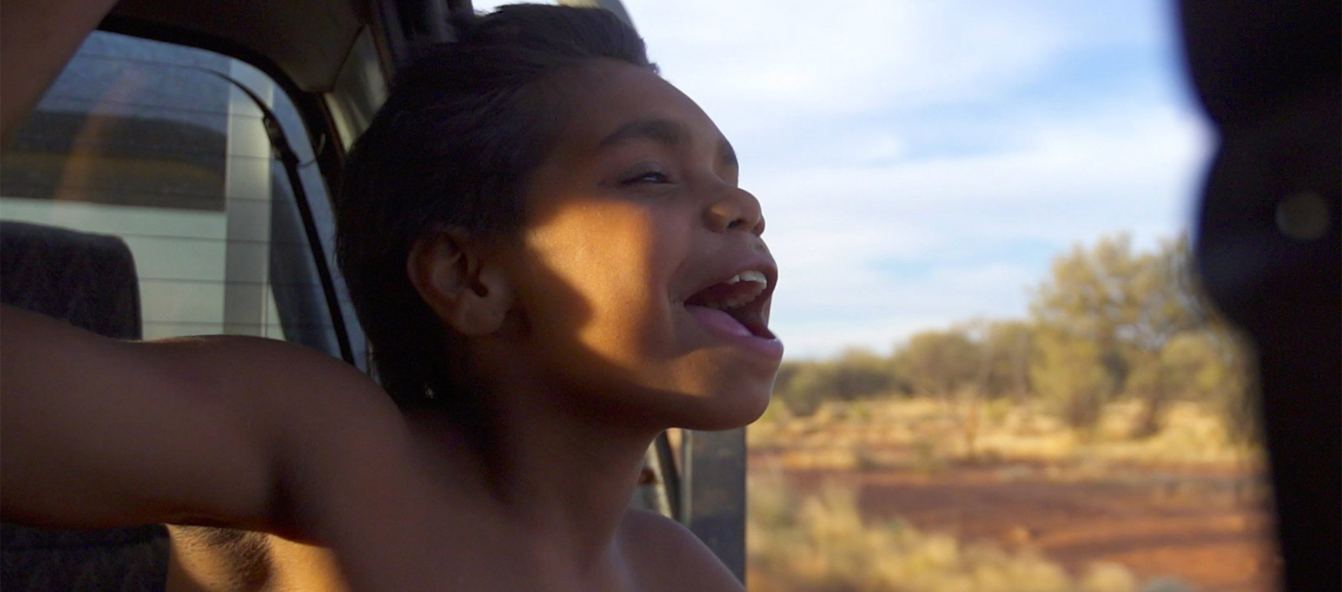 An aboriginal boy leans his head outside the window of a moving car