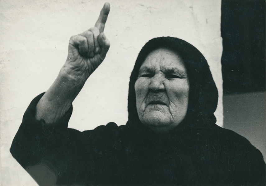 A black and white still of an elderly woman with her index finger raised in toward the sky