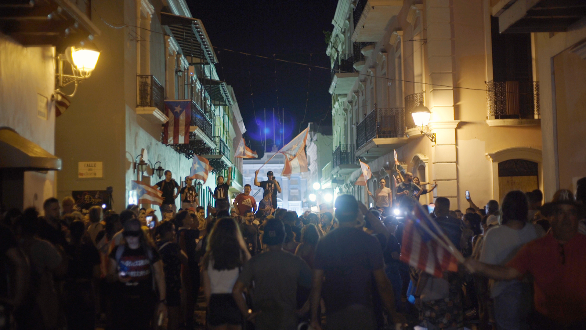 A crowd of people gather in the street, in the center of the frame is a person waving a flag