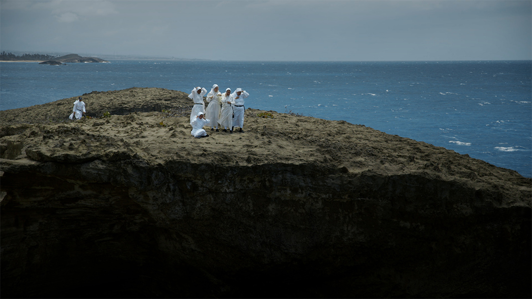 A group of people wearing white robes and head coverings stand near an edge of a cliff by a sea