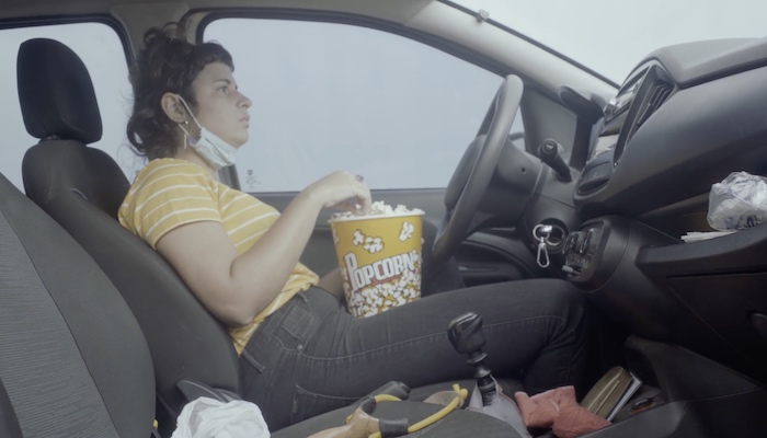 A woman sitting in the driver's seat of her car eating from a tub of popcorn in a scene from the Cinetracts '20 short by Gabriel Mascaro