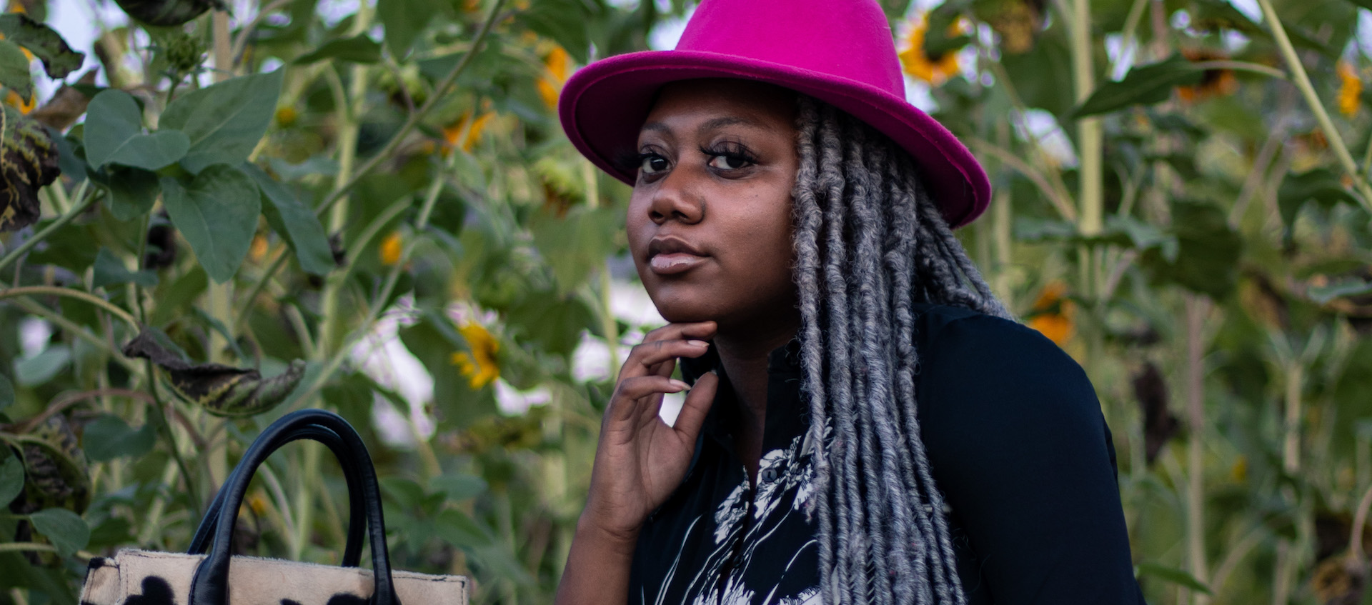 Artist Deijah Archie-Davis stands outside in a bright pink fedora in front of a field of sunflowers