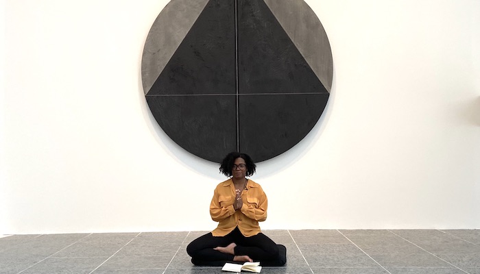Monique McCrystal sits on the tile floor of the Wexner Center for the Arts galleries with her hands in prayer pose, underneath a round abstract painting in black and gray by Torkwase Dyson