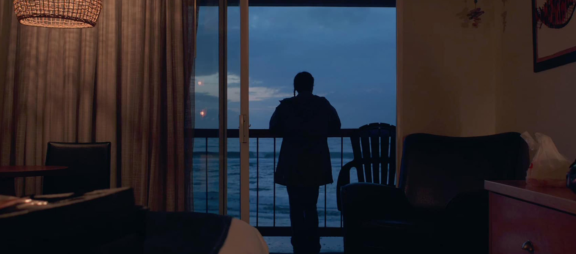 A person, seen from behind and in silhouette, looks out over the ocean on the balcony of a small room