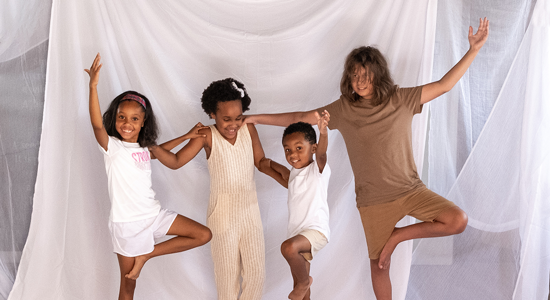  Photo of four children, all of whom have varying shades of brown skin and hair, standing in yoga poses in front of a background of white fabric