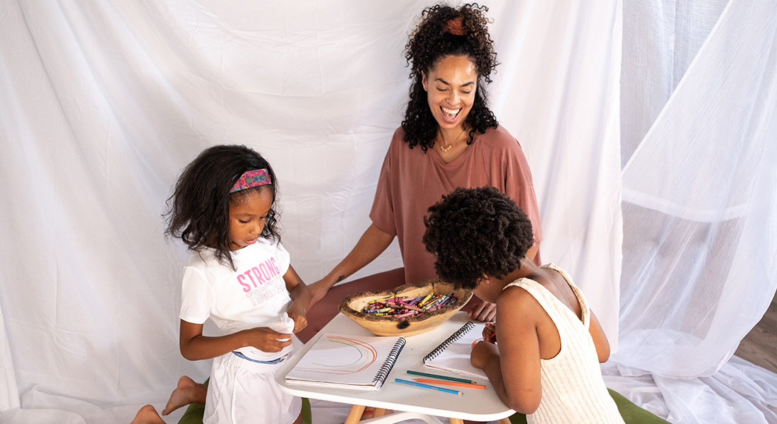 Photo of an instructor and young children, all of whom have varying shades of brown skin and hair, kneeling at a small, white table and drawing. Behind them are drapes of white fabric.