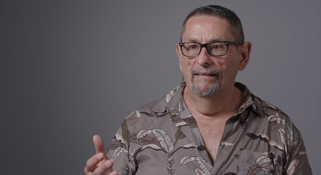 A person wearing dark glasses and a goatee sits in front of a gray wall. They wear a button down shirt, the top button is undonw