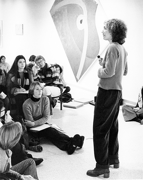 A black-and-white image of Elizabeth Murray standing in a gallery in front of several students seated to her left; a painting hangs on the far wall.