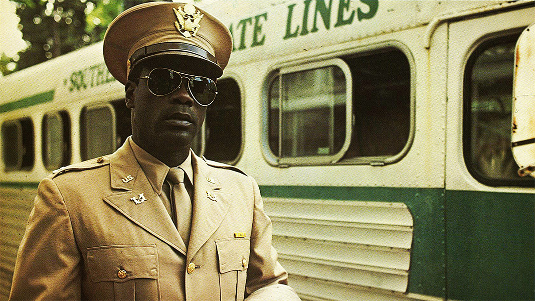 A man in beige military dress and sunglasses stands in front of a bus