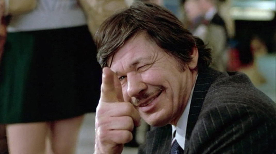 Charles Bronson stares ahead, blinking one eye and pointing his finger in the shape of a gun