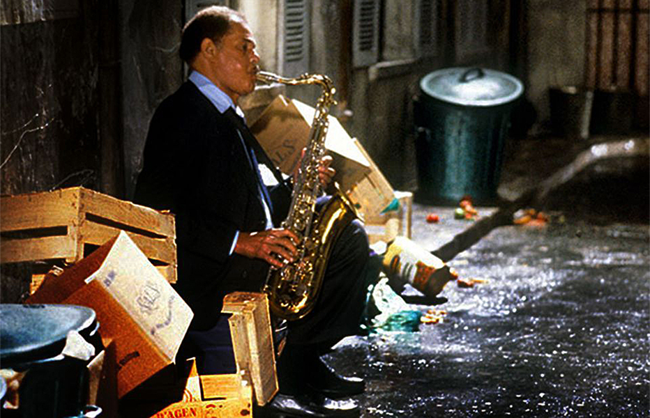 A man, seated in an alley plays a saxophone. He is surrounded by boxes and garbage.